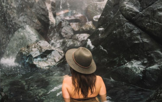 woman wearing wide brim hat facing wall of rocks while sitting in hot spring pool