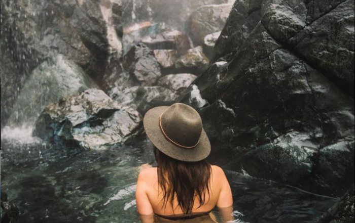 woman wearing wide brim hat facing wall of rocks while sitting in hot spring pool